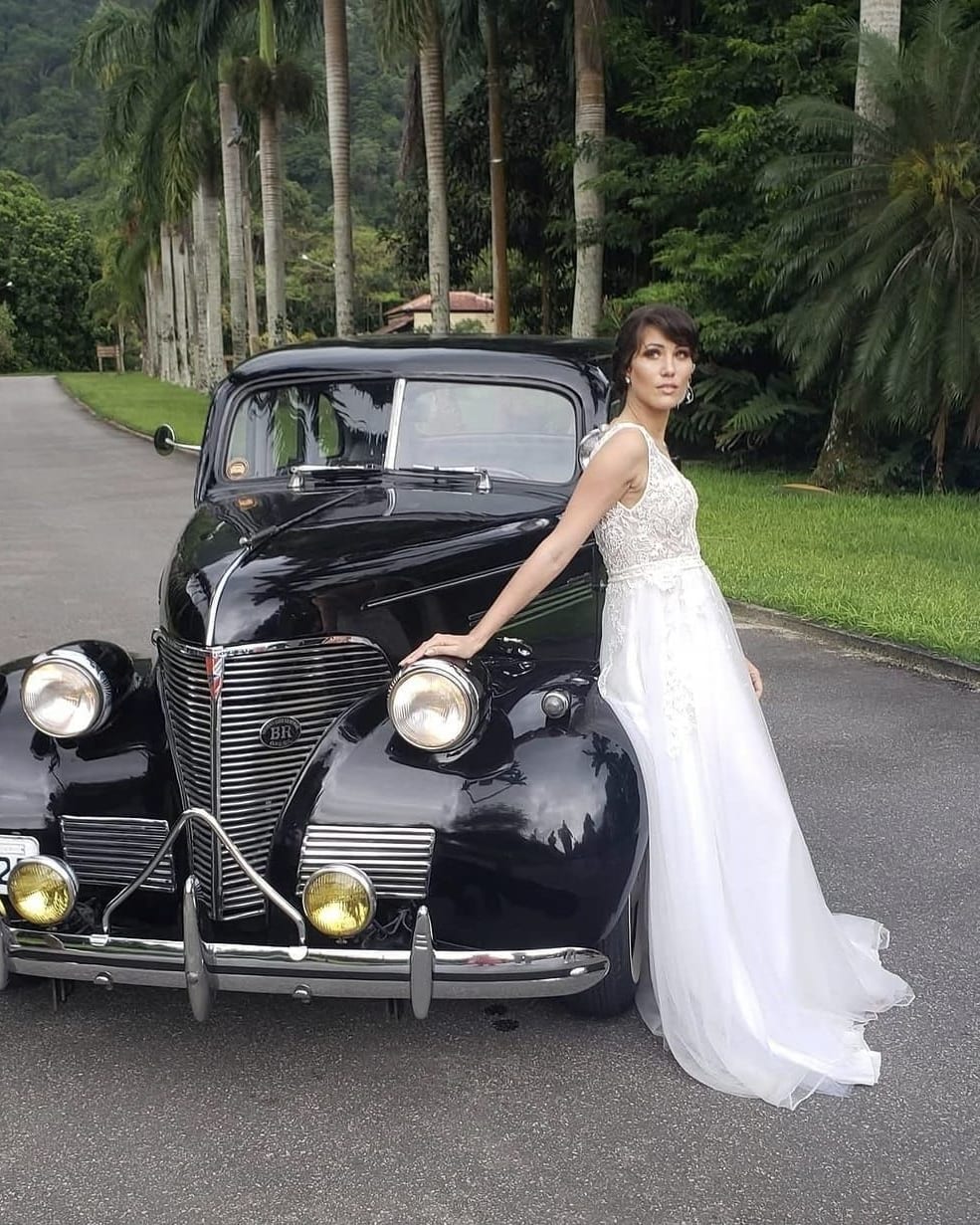 Noiva com penteado preso e maquiagem natural ao lado de um carro antigo